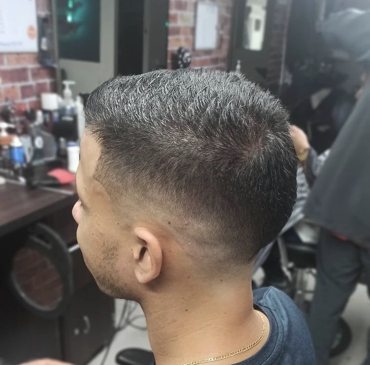 Hombre de perfil con corte corto y degradado bajo, en una barbería con paredes de ladrillo