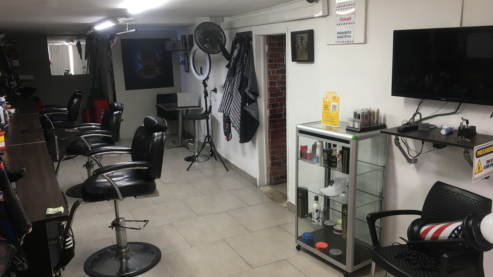 Salón con tres sillas de barbero negras, vitrina de productos, televisor y aro de luz